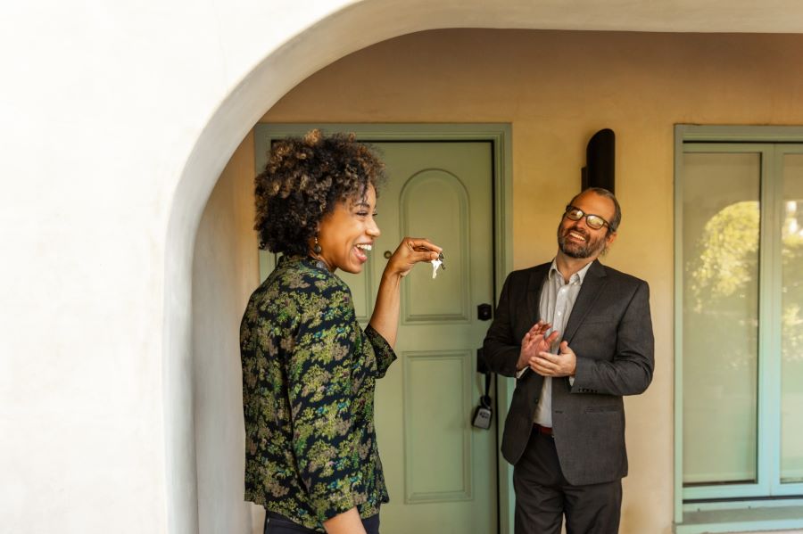 A man and a woman standing outside of a house. The woman excitedly holds up the keys to the front door.