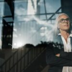 Confident woman looking out a window with arms crossed, reflecting on strategic decisions.