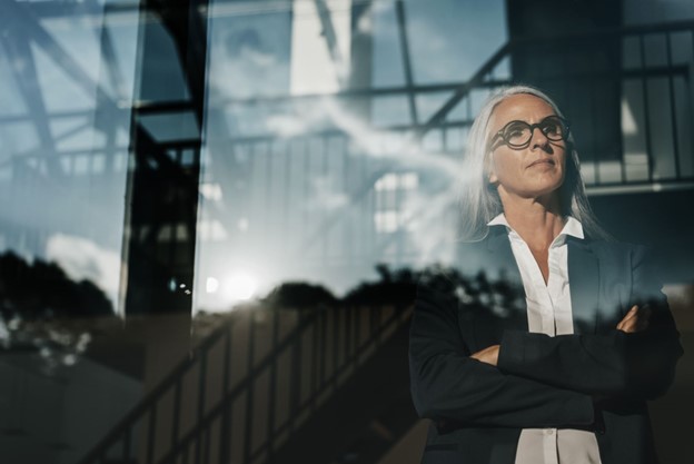 Confident woman looking out a window with arms crossed, reflecting on strategic decisions.